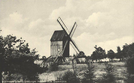 Winser Bockwindmühle Ein Schwarz-Weiß-Foto einer alten Windmühle in einer ländlichen Landschaft. Die Windmühle hat große Flügel und steht auf einem hölzernen Unterbau. Um die Mühle herum sind Bäume und Büsche zu sehen. Im Hintergrund sind weitere Gebäude und eine Gruppe von Menschen erkennbar.