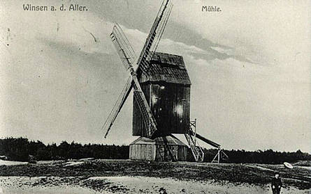 Winser Bockwindühle Ein Schwarz-Weiß-Foto einer Windmühle bei Winsen an der Aller. Die Mühle steht auf einem offenen Feld und hat große Flügel sowie eine hölzerne Struktur. Im Hintergrund ist eine Baumlinie zu sehen. Am rechten Bildrand steht eine Person, die in Richtung der Mühle schaut.