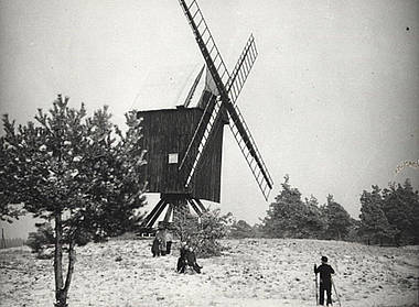 Winser Bockwindmühle Ein Schwarz-Weiß-Foto einer alten Bockwindmühle auf einem Hügel, umgeben von Bäumen. Vor der Mühle stehen mehrere Personen, die das Gebäude betrachten. Die Windmühle hat große Flügel.