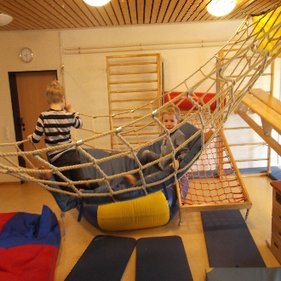 Kindertagesstätte Hinteres Sandfeld Zwei Kinder spielen in einem Raum mit Klettergerüsten und Hängematten aus Seilen. Ein Kind sitzt in der Hängematte und lächelt, während das andere Kind daneben steht. Der Raum hat eine Holzdecke und an den Wänden sind Kletterleitern befestigt. Auf dem Boden liegen weiche Matten in verschiedenen Farben.