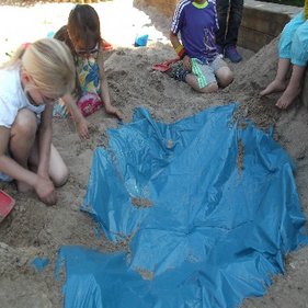 Kindertagesstätte Hinteres Sandfeld Mehrere Kinder spielen im Sandkasten und graben mit den Händen im Sand. Eine blaue Plane ist teilweise im Sand vergraben. Die Kinder tragen bunte Kleidung und sind konzentriert bei ihrer Tätigkeit.