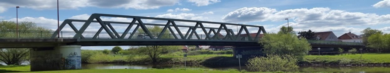 Wirtschaftsförderung Grüne Stahlbrücke über einen Fluss, umgeben von Wiesen und Bäumen, blauer Himmel mit Wolken.