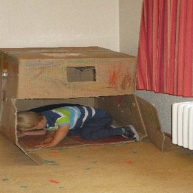 Kindertagesstätte Hinteres Sandfeld Ein Kind kriecht in eine große, bemalte Pappkiste, die als Spielhaus dient. Der Raum hat helle Wände und einen Heizkörper unter einem Fenster mit roten Vorhängen. Auf dem Boden liegen bunte Papierstücke verstreut.
