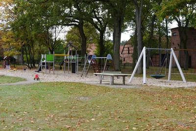 Spielplatz Stedden – Vorschaubild Blick über den Spielplatz in Stedden: Sandfläche mit Klettergerüst und Nestschaukel unter Bäumen, davor eine Rasenfläche.