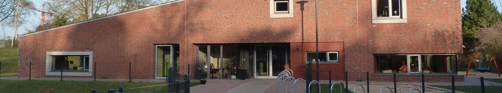 Kindertagesstätte Kleines Neues Land Ein modernes Gebäude mit einer Fassade aus roten Ziegeln und großen Fenstern. Vor dem Gebäude befindet sich ein gepflasterter Weg und ein umzäuntes Areal mit Fahrradständern. Das Gebäude hat ein schräges Dach und mehrere Eingänge. Im Hintergrund sind Bäume und ein klarer blauer Himmel zu sehen.