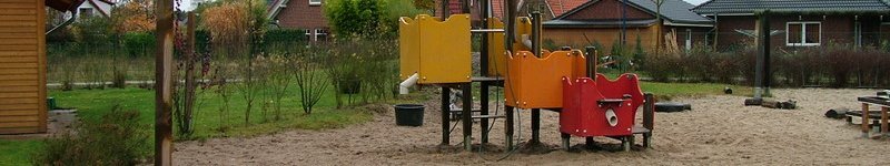 Kindertagesstätte Hinteres Sandfeld Ein Spielplatz mit einem bunten Klettergerüst auf einer großen Sandfläche. Im Hintergrund sind mehrere Wohnhäuser mit roten und dunklen Dächern zu sehen. Der Spielplatz ist von Grünflächen und Büschen umgeben, und es ist ein bewölkter Tag.