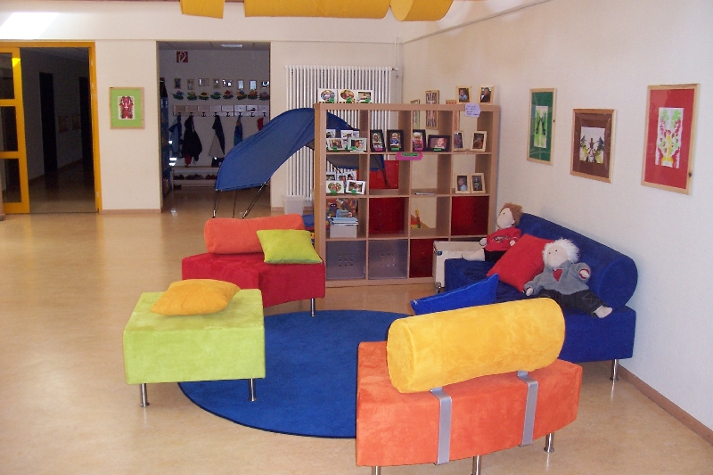 Kindertagesstätte Hinteres Sandfeld Ein heller Raum mit bunten Möbeln, darunter Sessel in Grün, Rot, Blau und Gelb. Die Möbel sind um einen runden blauen Teppich angeordnet. Im Hintergrund stehen Regale mit Büchern und Spielzeug. An der Wand hängen bunte Bilder. Eine Tür führt zu einem weiteren Raum, in dem eine Garderobe mit Kindersachen zu sehen ist.