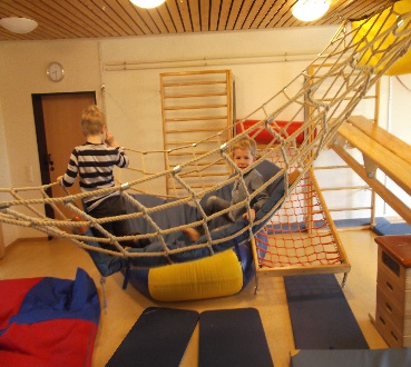 Kindertagesstätte Hinteres Sandfeld Zwei Kinder spielen in einem Raum mit Klettergerüsten und Hängematten aus Seilen. Ein Kind sitzt in der Hängematte und lächelt, während das andere Kind daneben steht. Der Raum hat eine Holzdecke und an den Wänden sind Kletterleitern befestigt. Auf dem Boden liegen weiche Matten in verschiedenen Farben.