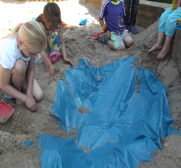 Kindertagesstätte Hinteres Sandfeld Mehrere Kinder spielen im Sandkasten und graben mit den Händen im Sand. Eine blaue Plane ist teilweise im Sand vergraben. Die Kinder tragen bunte Kleidung und sind konzentriert bei ihrer Tätigkeit.