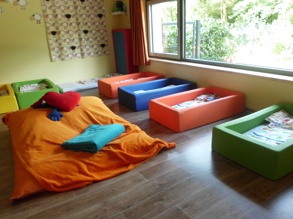 Kindertagesstätte Kleines Neues Land Ein Schlafraum für Kinder mit mehreren bunten Betten auf dem Boden. Die Betten sind in verschiedenen Farben, darunter Grün, Blau, Orange und Gelb. Ein großes Fenster mit Blick auf den Garten lässt viel Licht herein. Auf einem großen orangefarbenen Bett liegen ein rotes Herz- und ein blaues Kissen. Die Wände sind hellgelb und mit einem Vorhang mit Schafmuster dekoriert.