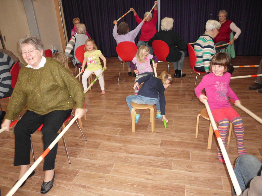 Kindertagesstätte Kleines Neues Land Eine Gruppe von Senioren und Kindern sitzt auf Stühlen und Hockern in einem Raum und führt gemeinsam Übungen mit langen Stäben durch. Die Senioren sitzen auf roten Stühlen, während die Kinder auf Hockern sitzen. Alle scheinen Freude an der Aktivität zu haben. Im Hintergrund hängt ein dunkler Vorhang.