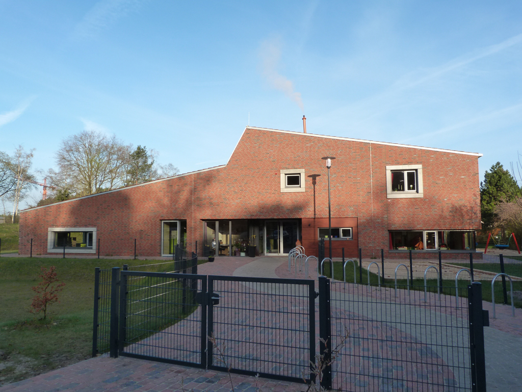 Kindertagesstätte Kleines Neues Land Ein modernes Gebäude mit einer Fassade aus roten Ziegeln und großen Fenstern. Vor dem Gebäude befindet sich ein gepflasterter Weg und ein umzäuntes Areal mit Fahrradständern. Das Gebäude hat ein schräges Dach und mehrere Eingänge. Im Hintergrund sind Bäume und ein klarer blauer Himmel zu sehen.