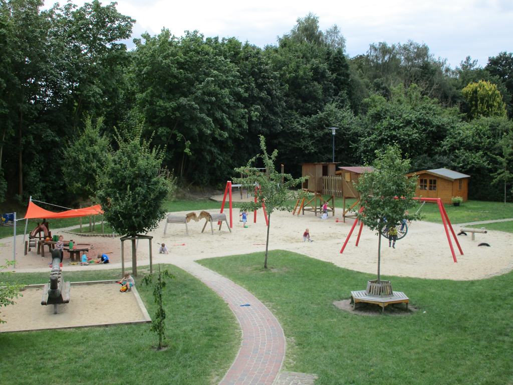 Kindertagesstätte Kleines Neues Land Ein großer Spielplatz mit verschiedenen Spielgeräten wie Schaukeln, Klettergerüsten und Sandkästen. Der Bereich ist mit Sand bedeckt und von Grünflächen und Bäumen umgeben. Einige Kinder spielen auf dem Spielplatz, während Erwachsene in der Nähe sitzen. Im Hintergrund ist ein kleines Holzhaus zu sehen, und die gesamte Szene ist von üppigem Grün und Bäumen eingerahmt.